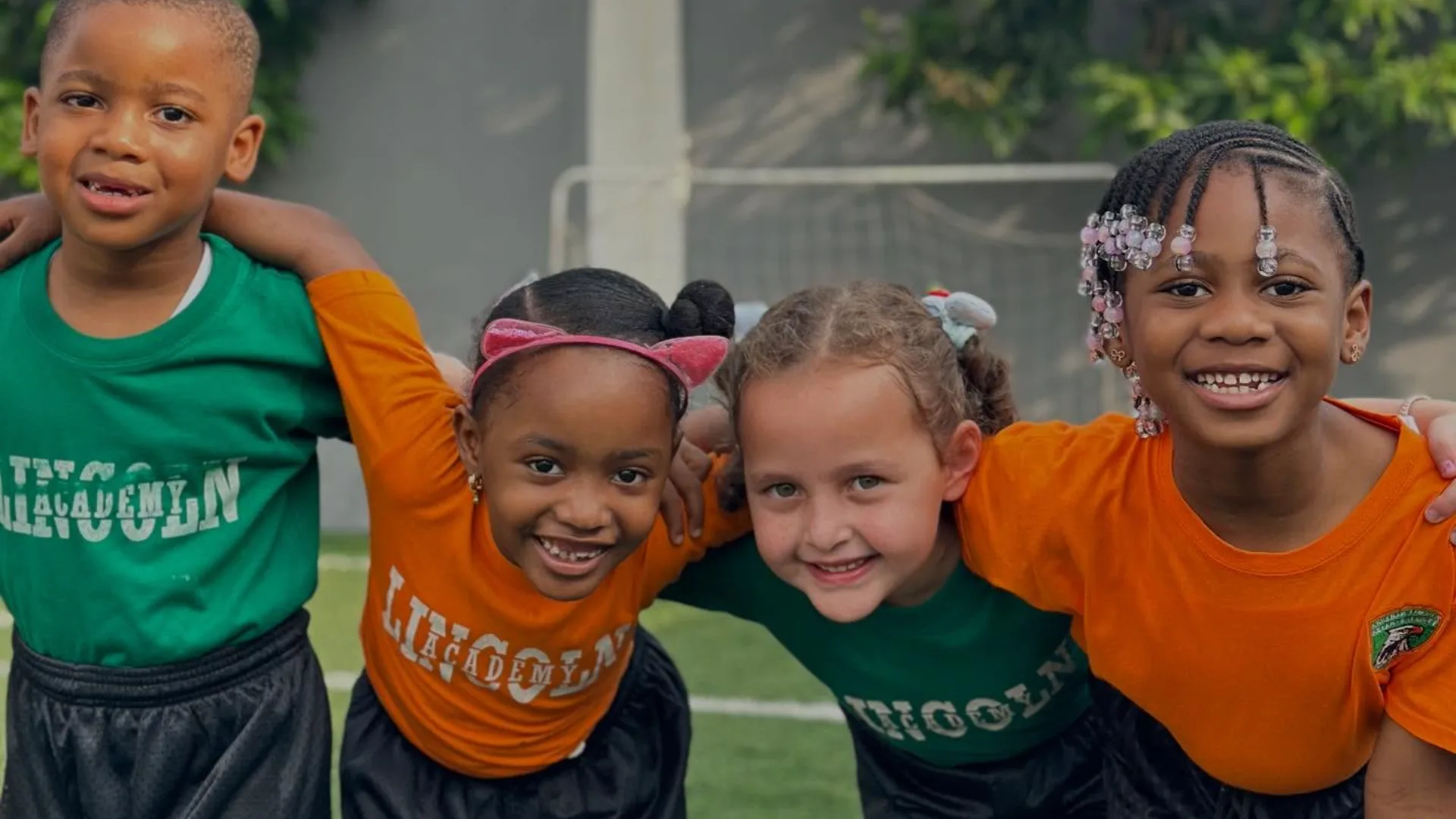 Four young children in green and orange sports shirts stand arm in arm on a sports pitch, smiling at the camera.