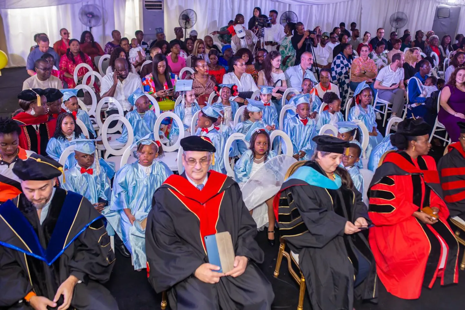 Children in pale blue graduation gowns and mortar boards sit with their families while staff in academic regalia take their places.